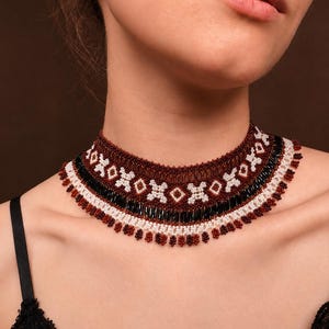 Puede incluir: Un collar tipo gargantilla de cuentas con un patrón geométrico en rojo oscuro, negro y blanco. El diseño consta de filas horizontales de pequeñas cuentas y cuentas tubulares alargadas, con un delicado fleco de pequeños racimos en el borde inferior.