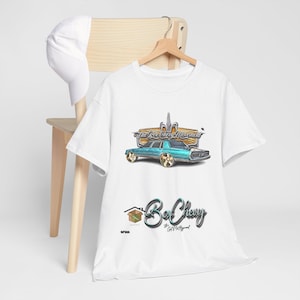 May include: White t-shirt with a teal car graphic and the text "The Box Chevy Movement" and "Be Chevy". A white baseball cap hangs on a wooden chair. The shirt is hanging on a wooden hanger.