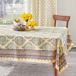 May include: A beige tablecloth with a detailed floral pattern covers a wooden table. The tablecloth has a border with a repeating design in shades of purple, orange, and green. Yellow curtains and a wooden chair are in the background. A vase of yellow flowers sits on the table.