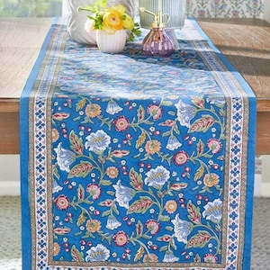 May include: A blue floral table runner with a detailed pattern of white, pink, and yellow flowers and green leaves. The runner has a border with a repeating geometric design. The runner is approximately 72 inches long.