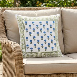 May include: A square throw pillow with a white background and a blue floral pattern. The pillow has a green border and is placed on a beige cushion on a wicker sofa. The sofa is outdoors in a garden setting.