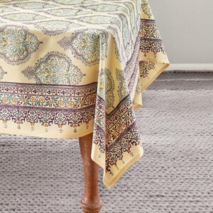 Puede incluir: Un mantel beige con un patr&oacute;n detallado y repetitivo en verde azulado, morado y naranja. El mantel est&aacute; drapeado sobre una mesa de madera, con la esquina de la tela colgando. El patr&oacute;n incluye elementos florales y geom&eacute;tricos.