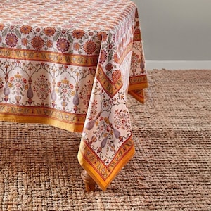 May include: A rectangular tablecloth with a floral pattern in shades of orange, red, and purple on a cream background. The tablecloth has a yellow border and is draped over a wooden table. The table is set on a woven rug.