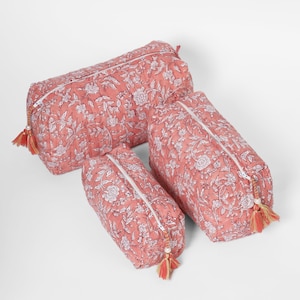 May include: A set of three quilted pouches in a dusty rose color with a white floral block print pattern. Each rectangular pouch features a white zipper closure accented with a decorative tassel pull containing small beads and multi-colored threads.