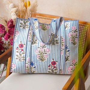 May include: A large tote bag with a floral pattern and blue stripes. The bag features pink and red flowers with green leaves. The handles and edges are trimmed with a blue and white striped fabric. The bag is sitting on a wooden chair.