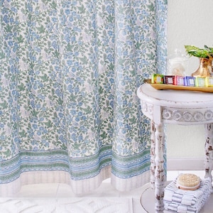 Op de afbeelding: Een witte badkamer met een bloemen douchegordijn, een witte badmat en een witte bijzettafel met een houten blad. De bijzettafel heeft een kleine houten kom met zeepjes.