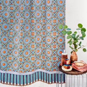 May include: A blue and orange shower curtain with a geometric pattern. The curtain has a white border with a blue and green stripe pattern. The curtain is hanging in a bathroom with a side table holding a brown vase with green leaves, a brown bottle, and a red and white patterned soap dispenser.
