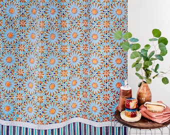 May include: A blue and orange shower curtain with a geometric pattern. The curtain has a white border with a blue and green stripe pattern. The curtain is hanging in a bathroom with a side table holding a brown vase with green leaves, a brown bottle, and a red and white patterned soap dispenser.