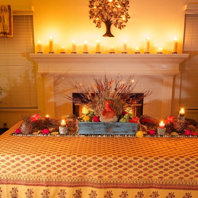 May include: A table with a yellow and orange patterned tablecloth. A blue wooden box with a fall floral arrangement is on the table. The arrangement includes a turkey figurine, fall leaves, and pine cones. There are candles on the table and a fireplace in the background.