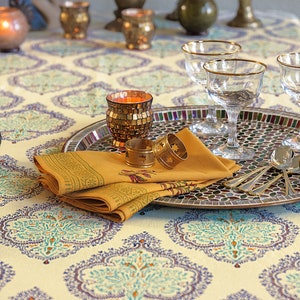 Puede incluir: Una mesa decorada con un mantel estampado en crema, turquesa y azul. Una bandeja con mosaicos contiene copas con borde dorado, servilletas mostaza dobladas y servilleteros dorados. Velas decorativas y piezas para servir completan el elegante arreglo.