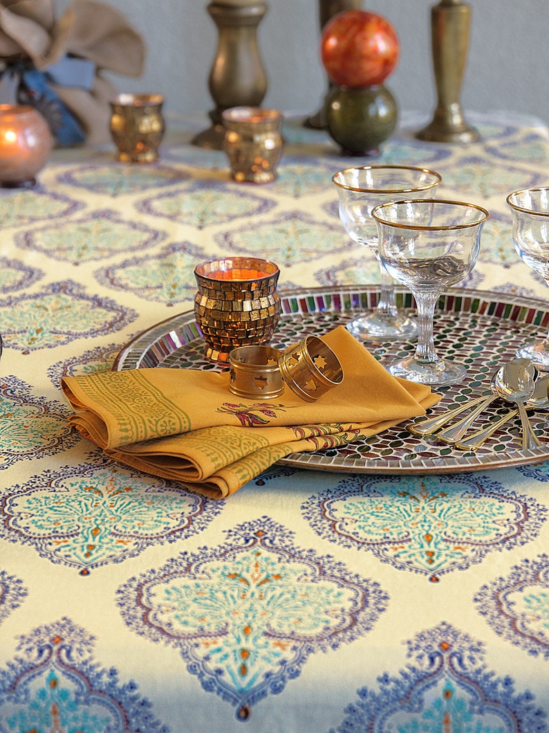 Puede incluir: Una mesa decorada con un mantel estampado en crema, turquesa y azul. Una bandeja con mosaicos contiene copas con borde dorado, servilletas mostaza dobladas y servilleteros dorados. Velas decorativas y piezas para servir completan el elegante arreglo.
