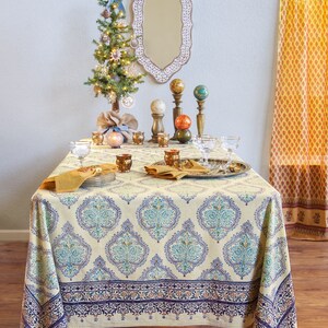 Puede incluir: Una mesa de comedor decorada para una ocasi&oacute;n festiva, con un mantel crema con un estampado floral azul y turquesa. La mesa est&aacute; adornada con cristaler&iacute;a, servilletas y candelabros decorativos. Un peque&ntilde;o &aacute;rbol de Navidad decorado y un espejo decorativo est&aacute;n en el fondo.