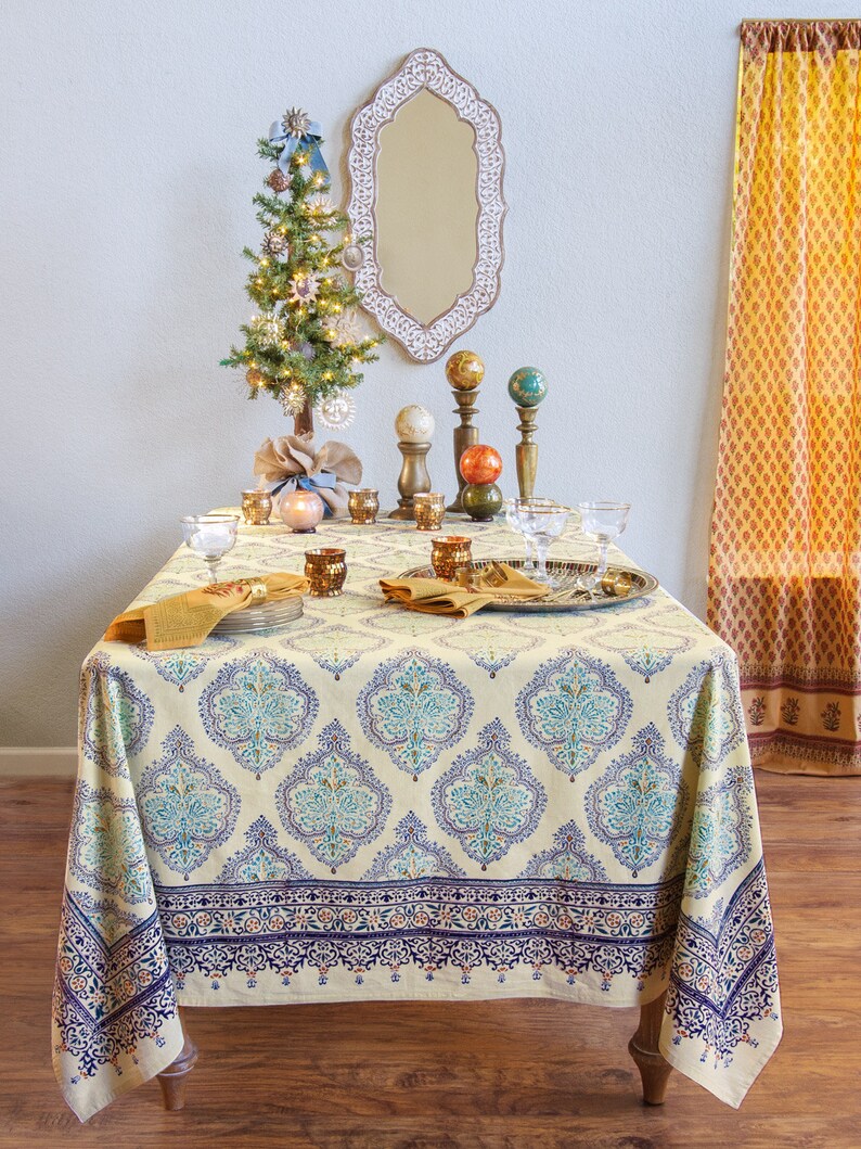 Puede incluir: Una mesa de comedor decorada para una ocasi&oacute;n festiva, con un mantel crema con un estampado floral azul y turquesa. La mesa est&aacute; adornada con cristaler&iacute;a, servilletas y candelabros decorativos. Un peque&ntilde;o &aacute;rbol de Navidad decorado y un espejo decorativo est&aacute;n en el fondo.