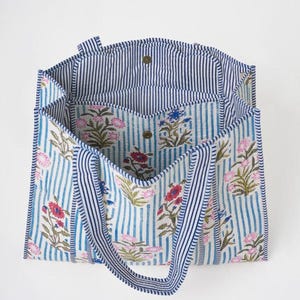 May include: A tote bag featuring a light blue and white vertical striped pattern with a floral print in shades of pink, red, and blue. The bag has two striped handles, a wide open top, and a central interior compartment with a snap button closure.