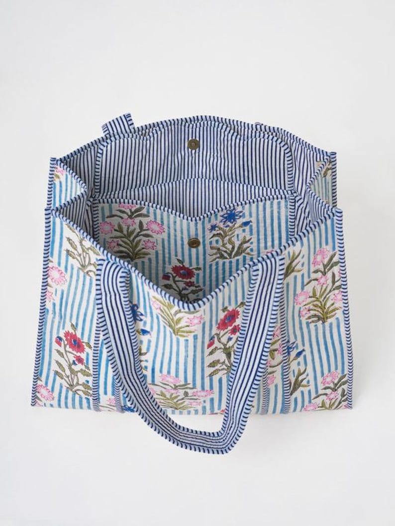 May include: A tote bag featuring a light blue and white vertical striped pattern with a floral print in shades of pink, red, and blue. The bag has two striped handles, a wide open top, and a central interior compartment with a snap button closure.