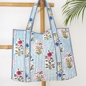 May include: A rectangular tote bag featuring a white and blue vertical striped pattern with a floral print in shades of pink, red, and blue. The bag has two matching striped fabric handles and is constructed with visible stitching along the edges.