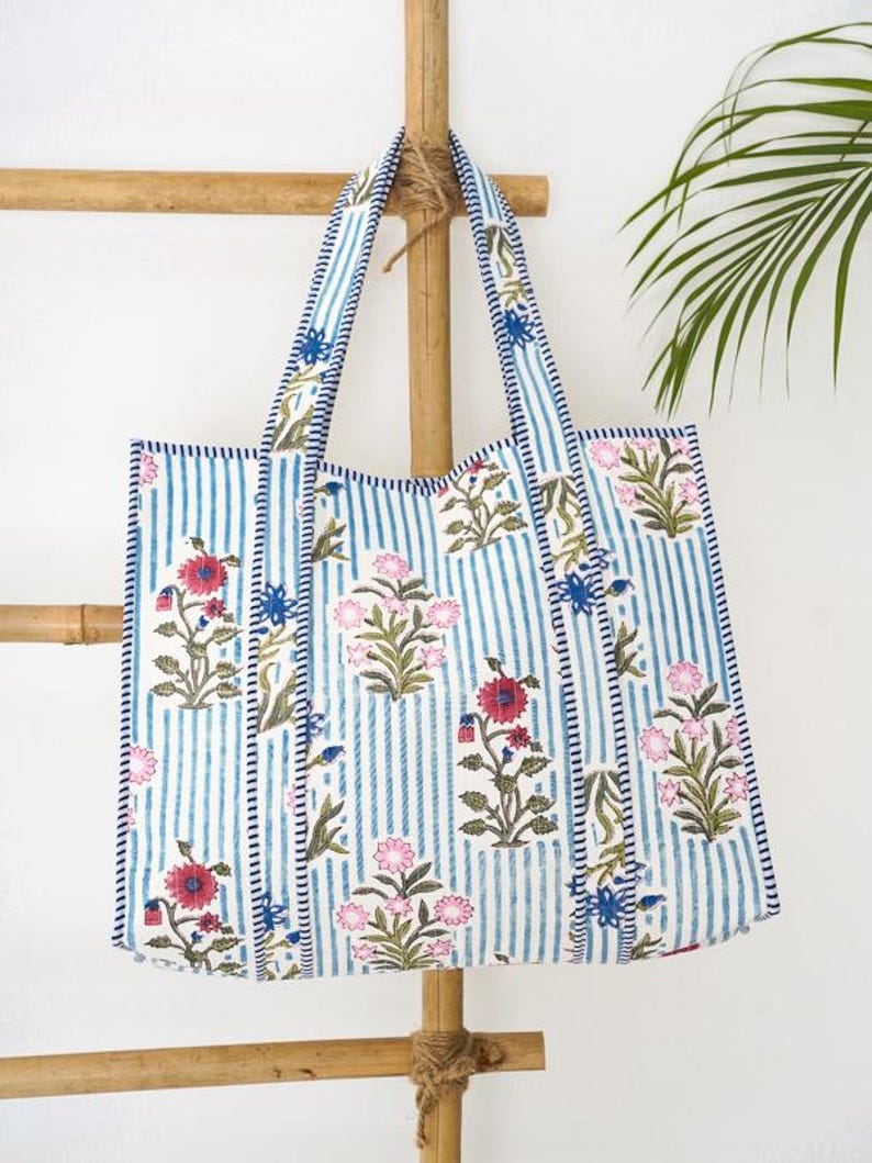 May include: A rectangular tote bag featuring a white and blue vertical striped pattern with a floral print in shades of pink, red, and blue. The bag has two matching striped fabric handles and is constructed with visible stitching along the edges.