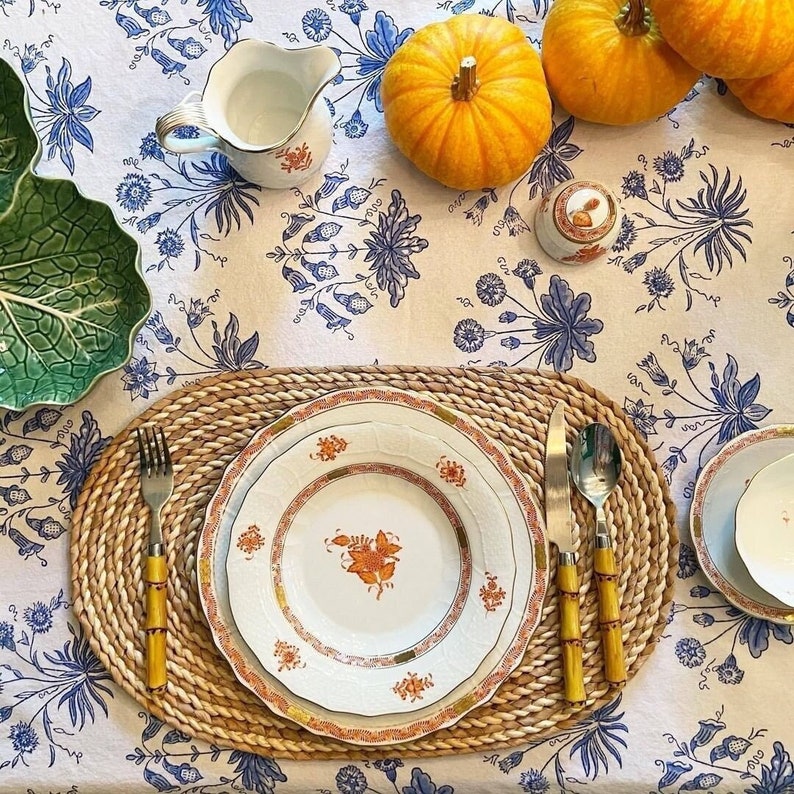 May include: A table setting with white plates featuring orange floral designs, bamboo-handled silverware, and a woven placemat. The table is covered with a white tablecloth with blue floral patterns. Pumpkins and serving dishes complete the autumnal scene.