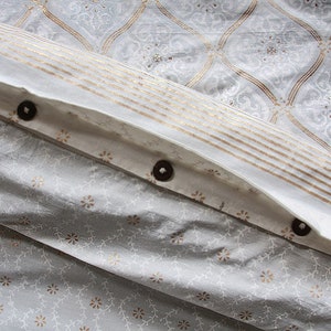 May include: White fabric with a gold and white pattern. The fabric has a fold with three brown buttons.