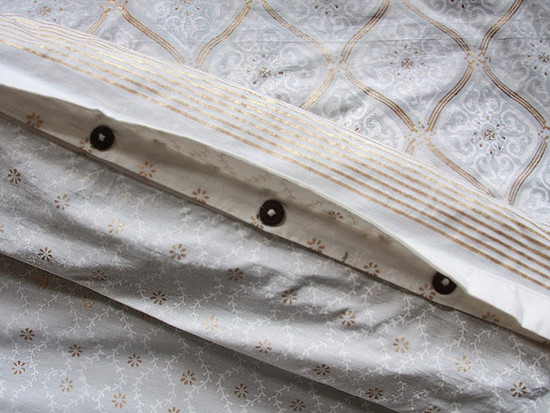 May include: White fabric with a gold and white pattern. The fabric has a fold with three brown buttons.