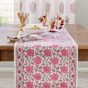 May include: A floral table runner with a pink and white pattern, set on a wooden table. The runner features a repeating design of large pink flowers and leaves. Matching napkins and a bowl are also on the table, along with desserts and breadsticks.