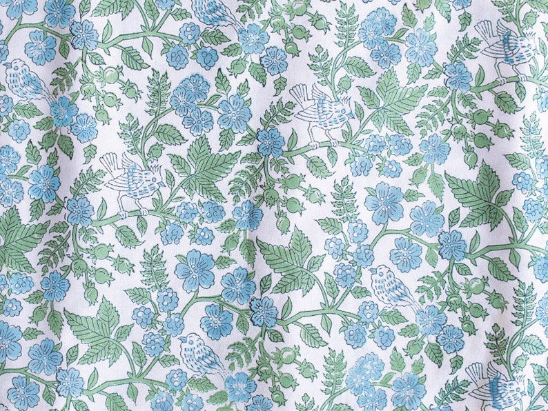 Op de afbeelding: Witte stof met een herhalend patroon van blauwe bloemen, groene bladeren en witte vogels. Het patroon is in traditionele stijl.
