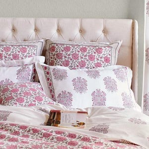 K&ouml;nnte beinhalten: Ein wei&szlig;es Bett mit einem gesteppten Kopfteil und einem wei&szlig;en Bettbezug mit rosafarbenem Blumenmuster. Auf dem Bett liegen mehrere Kissen mit rosafarbenen Blumenmustern. Ein Buch liegt auf dem Bett.