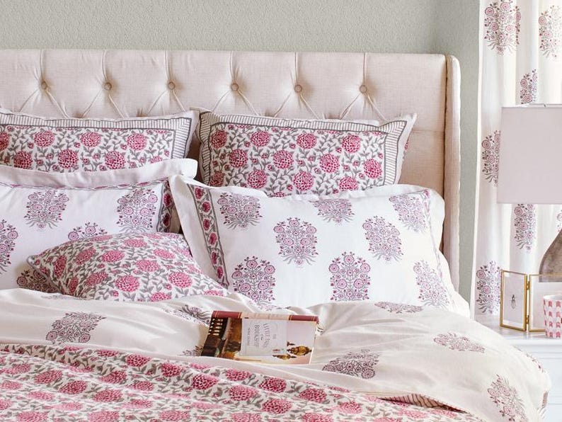K&ouml;nnte beinhalten: Ein wei&szlig;es Bett mit einem gesteppten Kopfteil und einem wei&szlig;en Bettbezug mit rosafarbenem Blumenmuster. Auf dem Bett liegen mehrere Kissen mit rosafarbenen Blumenmustern. Ein Buch liegt auf dem Bett.