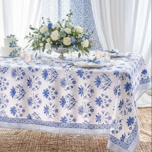 May include: A dining table set for a special occasion. The white tablecloth features a blue floral pattern. A centerpiece of white and blue flowers sits in a silver vase. Matching cake and curtains complete the elegant setting.