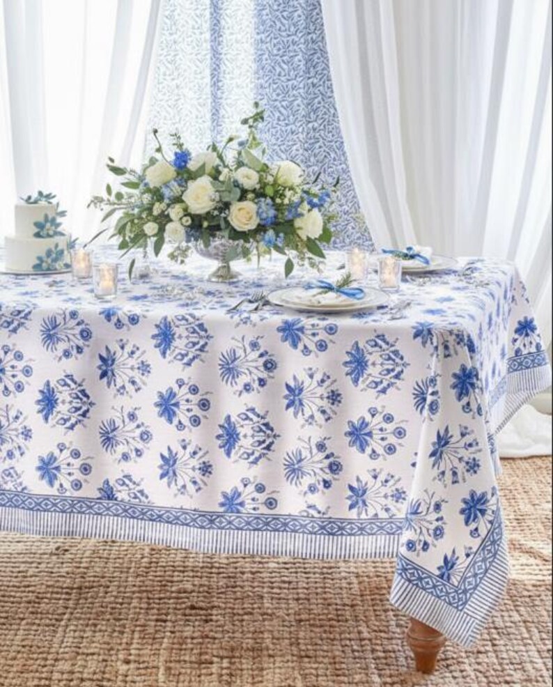May include: A dining table set for a special occasion. The white tablecloth features a blue floral pattern. A centerpiece of white and blue flowers sits in a silver vase. Matching cake and curtains complete the elegant setting.