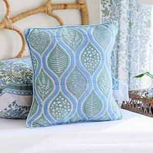 May include: Square decorative pillow with a light blue border and a repeating pattern of green leaf-like shapes on a white background. The design is framed by a wavy light blue border. The pillow is on a white bed.