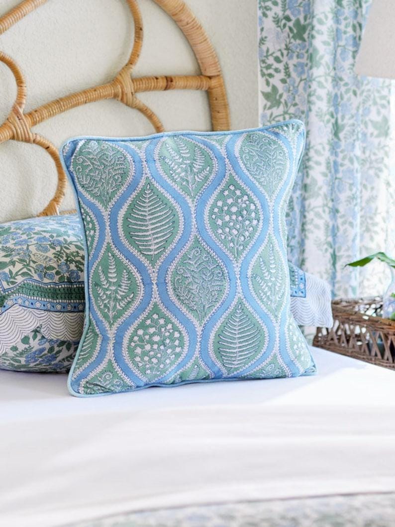 May include: Square decorative pillow with a light blue border and a repeating pattern of green leaf-like shapes on a white background. The design is framed by a wavy light blue border. The pillow is on a white bed.
