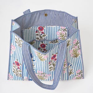 May include: A tote bag featuring a light blue and white vertical striped pattern with a floral print in shades of pink, red, and green. The bag has two matching striped handles, a wide open top, and a single gold-toned snap button closure.