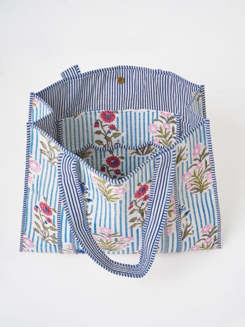 May include: A tote bag featuring a light blue and white vertical striped pattern with a floral print in shades of pink, red, and green. The bag has two matching striped handles, a wide open top, and a single gold-toned snap button closure.