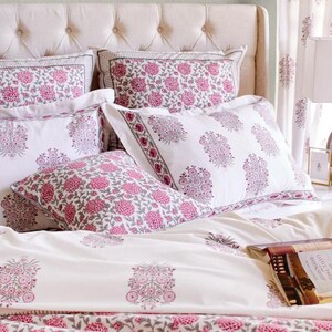 K&ouml;nnte beinhalten: Ein wei&szlig;es Bett mit einem rosafarbenen, floral gemusterten Bettbezug und Kissen. Das Bett ist mit einem wei&szlig;en Kopfteil und einem wei&szlig;en Nachttisch mit einer Lampe und einem Buch darauf ausgestattet.