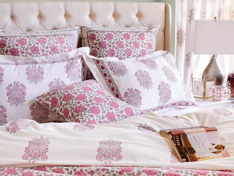 K&ouml;nnte beinhalten: Ein wei&szlig;es Bett mit einem rosafarbenen, floral gemusterten Bettbezug und Kissen. Das Bett ist mit einem wei&szlig;en Kopfteil und einem wei&szlig;en Nachttisch mit einer Lampe und einem Buch darauf ausgestattet.