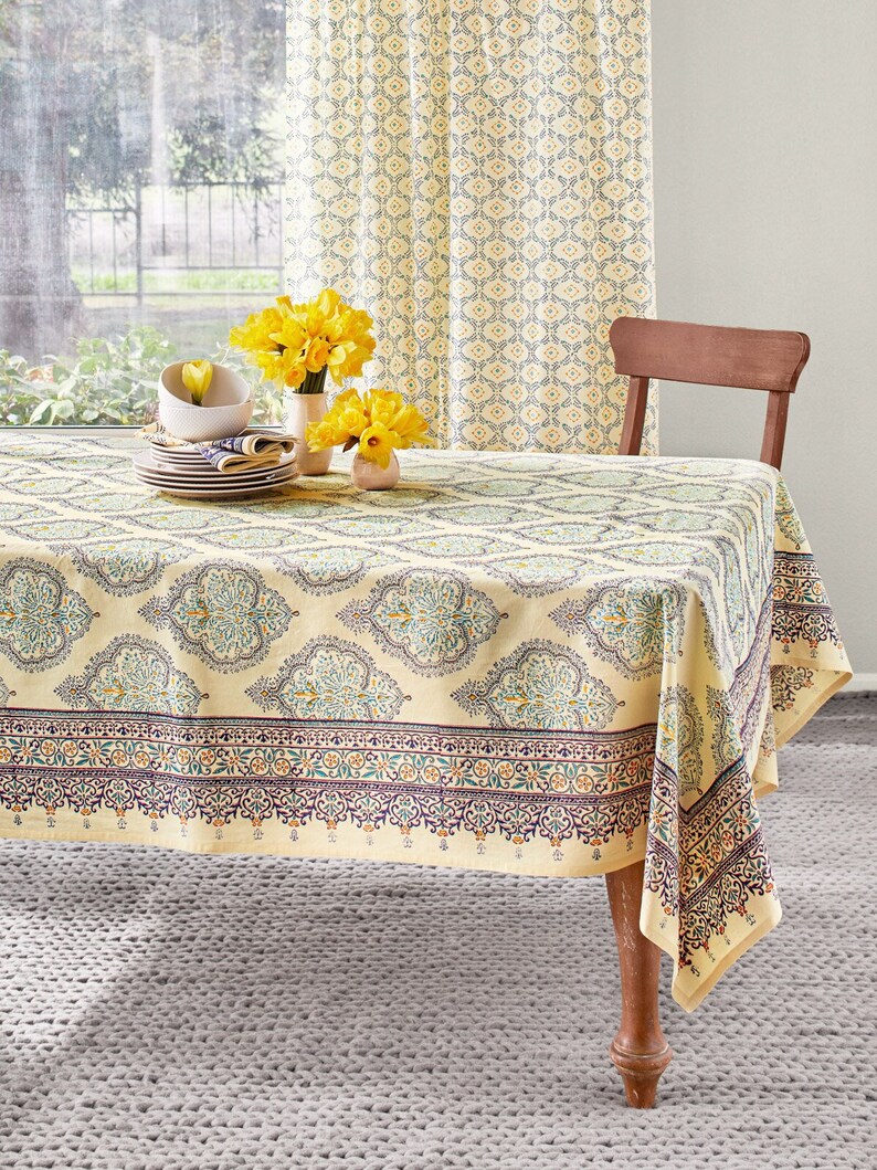 May include: A beige tablecloth with a detailed floral pattern covers a wooden table. The tablecloth has a border with a repeating design in shades of purple, orange, and green. Yellow curtains and a wooden chair are in the background. A vase of yellow flowers sits on the table.