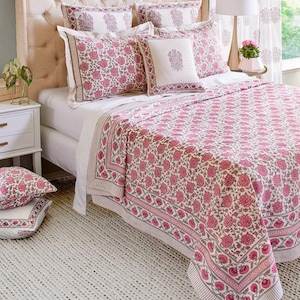 May include: A bed with a tufted headboard is covered with a floral patterned quilt and matching pillows. The quilt and pillows feature a pink floral design on a white background, with a gray border. A white rug is on the floor.