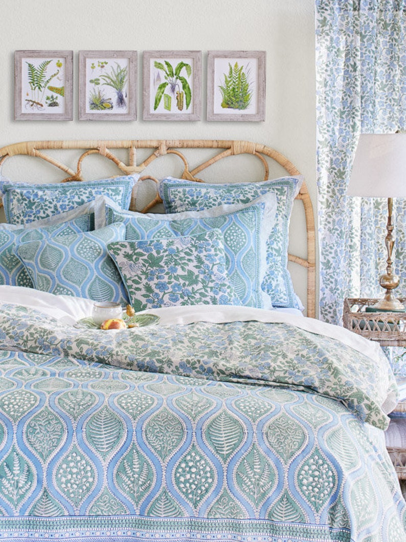 May include: A bedroom scene featuring a bed with a woven headboard and bedding in shades of blue and green. The bedding has a botanical pattern. Above the bed are four framed botanical prints. A matching curtain and a lamp complete the decor.