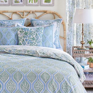 May include: A bed with a woven headboard and botanical-themed bedding. The bedding features a blue and green leaf pattern, complemented by matching pillows and a duvet cover. Four framed botanical prints hang above the bed, and a patterned curtain is visible.