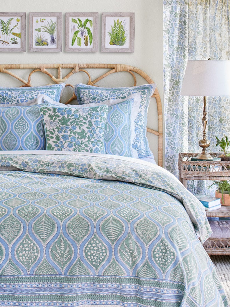 May include: A bed with a woven headboard and botanical-themed bedding. The bedding features a blue and green leaf pattern, complemented by matching pillows and a duvet cover. Four framed botanical prints hang above the bed, and a patterned curtain is visible.
