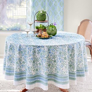 Op de afbeelding: Een wit tafelkleed met een blauw en groen bloemenpatroon. Het tafelkleed heeft een rand met een herhalend patroon van blauwe en groene strepen. Het tafelkleed ligt op een ronde tafel met een houten onderstel. Er staan twee wijnglazen en een bord met eten op de tafel.