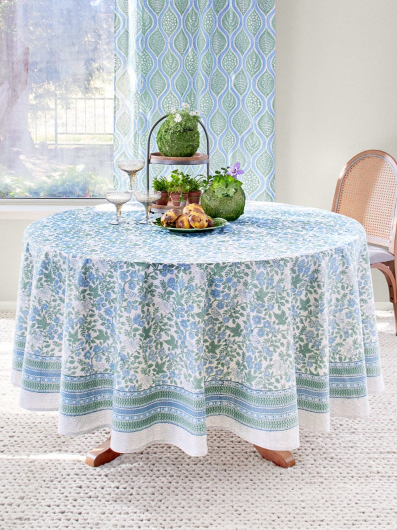 Op de afbeelding: Een wit tafelkleed met een blauw en groen bloemenpatroon. Het tafelkleed heeft een rand met een herhalend patroon van blauwe en groene strepen. Het tafelkleed ligt op een ronde tafel met een houten onderstel. Er staan twee wijnglazen en een bord met eten op de tafel.