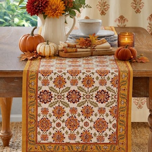 May include: A rectangular table runner featuring a repeating floral pattern in shades of orange, yellow, deep red, and green on a cream background, bordered by a solid mustard yellow band with a decorative vine motif along the edges.