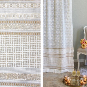 May include: White and gold patterned fabric, possibly a curtain or tablecloth, with repeating floral and dot designs. The fabric is draped, with a chair and flowers in the background. A tray with candles and champagne glasses is in the foreground.