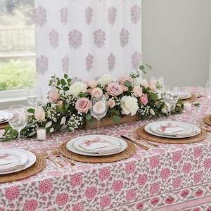 May include: A beautifully set table with a floral centerpiece in a wooden box. The table is covered with a pink floral tablecloth, and each place setting includes a plate, napkin, and wine glass. The background features a floral patterned curtain.