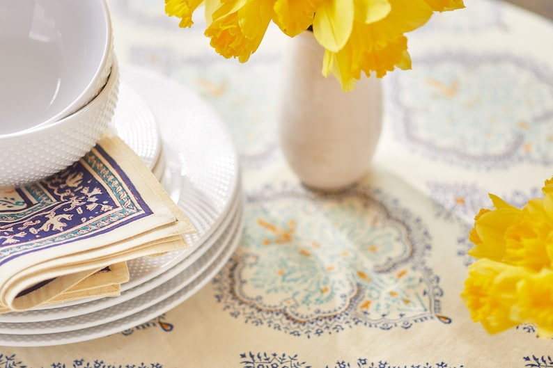 Puede incluir: Una pila de platos blancos y un cuenco con superficie texturizada se encuentran junto a servilletas dobladas con un estampado azul y turquesa. Un peque&ntilde;o jarr&oacute;n blanco contiene narcisos amarillos. La mesa est&aacute; cubierta con un mantel color crema con un dise&ntilde;o azul y naranja.
