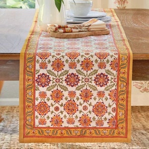 May include: A white table runner with a floral pattern in shades of orange, red, and purple. The runner has a gold border with a vine design. The runner is on a wooden table.