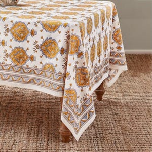 May include: A white tablecloth with a repeating pattern of yellow and blue paisley-like designs. The tablecloth has a border with a gray and blue geometric pattern. The table is set on a brown woven rug.