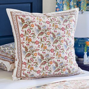 May include: A square pillow with a white background and a colorful floral pattern. The pattern includes red, blue, yellow, and green flowers and leaves. The pillow has a decorative border and is placed on a bed with a matching floral patterned bedspread.